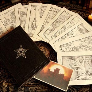 May include: A tarot card deck with a black book featuring a white pentagram. The cards are illustrated with black and white drawings. A small card with a sunset scene is also present. Lit candles and other objects are in the background.