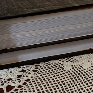 以下が含まれることがあります： 黒い表紙と白いページを持つ2冊のハードカバーの本が積み重ねられています。それらは、花の模様の白いレースのドイリーの上に置かれています。本の端が見え、個々のページが表示されています。読書や執筆に適しています。