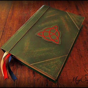 Puede incluir: Un diario artesanal de cuero verde con un emblema de nudo celta rojo. El libro tiene un acabado envejecido y presenta tres cintas de colores como marcadores. El diario se muestra sobre una superficie de madera.
