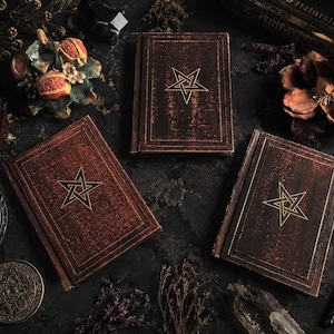 Könnte beinhalten: Drei Bücher im antiken Stil, braun ledergebunden, mit goldenen Pentagramm-Designs. Die Bücher sind auf einer dunklen Oberfläche mit Kerzen, Trockenblumen und einem dekorativen Schlüssel angeordnet. Die Gesamtästhetik ist gotisch und mystisch.