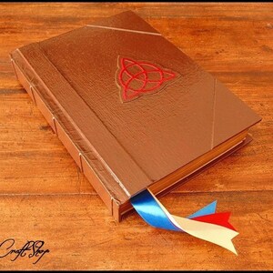 Puede incluir: Un libro encuadernado en cuero marr&oacute;n con un dise&ntilde;o de nudo celta rojo y un marcap&aacute;ginas de cinta azul, blanca y roja. El libro tiene un brillo met&aacute;lico y est&aacute; sobre una superficie de madera. El texto "MagiCraft Shop" est&aacute; abajo.