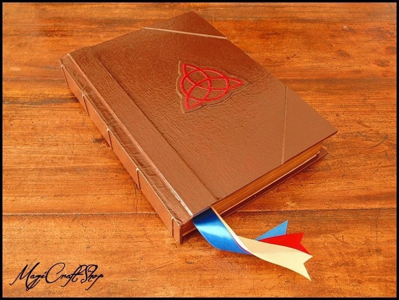 Puede incluir: Un libro encuadernado en cuero marr&oacute;n con un dise&ntilde;o de nudo celta rojo y un marcap&aacute;ginas de cinta azul, blanca y roja. El libro tiene un brillo met&aacute;lico y est&aacute; sobre una superficie de madera. El texto "MagiCraft Shop" est&aacute; abajo.