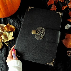 May include: A black leather-bound journal with a metal lock and keyhole, decorated with ornate brass corners. The journal is lying on a black surface with autumn leaves and a small crystal.