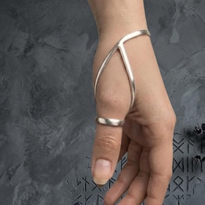 May include: A silver-toned hand ornament featuring a smooth, curved metal band that wraps around the thumb and extends upward to encircle the wrist. The piece consists of a simple ring base connected to a Y-shaped structure that rests on the back of the hand.