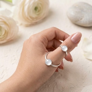 May include: Two silver rings featuring smooth, round, translucent white cabochon stones. One ring is worn on the thumb and the other on the index finger, both showcasing a minimalist metal band design with a polished finish and a clear, milky gemstone.