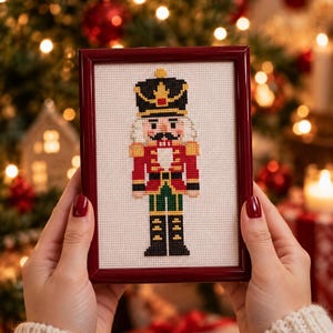 May include: A cross-stitch embroidery of a nutcracker soldier in a dark red, green, and black uniform with gold accents, displayed in a dark red rectangular frame against a white fabric background with a visible grid pattern.