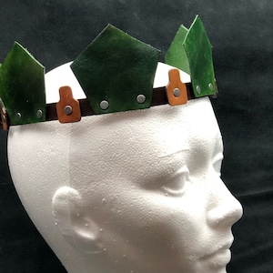 May include: A green leather crown with brown leather accents and silver metal rivets. The crown is worn on a white mannequin head.