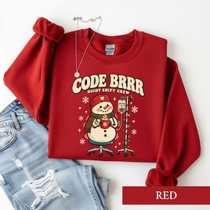 May include: A red long-sleeve sweatshirt featuring a graphic of a snowman wearing a red hat and green scarf, holding a mug of cocoa. The text reads CODE BRRR NIGHT SHIFT CREW. The snowman is sitting on a stool next to an IV pole with lights.
