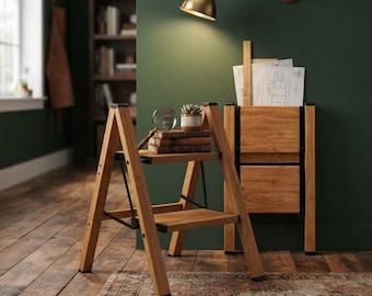May include: A light brown wooden step stool with two tiers and black metal hinges. On the top step sits a small potted succulent, a magnifying glass, and a stack of three brown books. Beside it stands a matching wooden storage unit.