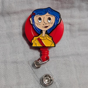 May include: A badge reel with a cartoon character design. The character has blue hair and a yellow jacket, set against a red background. The reel includes a clear plastic strap and a metal clip.