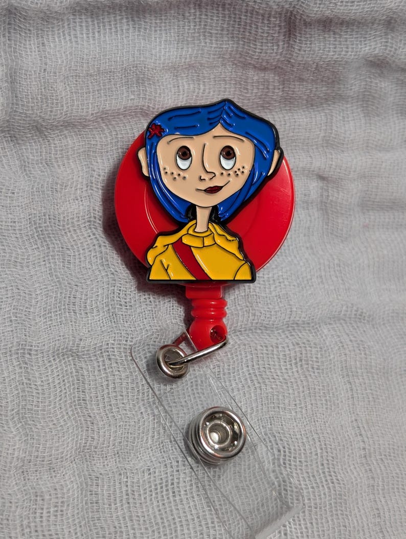 May include: A badge reel with a cartoon character design. The character has blue hair and a yellow jacket, set against a red background. The reel includes a clear plastic strap and a metal clip.