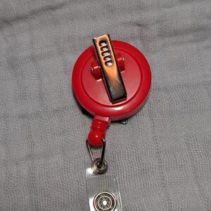 May include: A red retractable badge reel with a silver metal clip and a clear plastic strap. The reel is round and has a small red button. The strap has a metal snap closure.