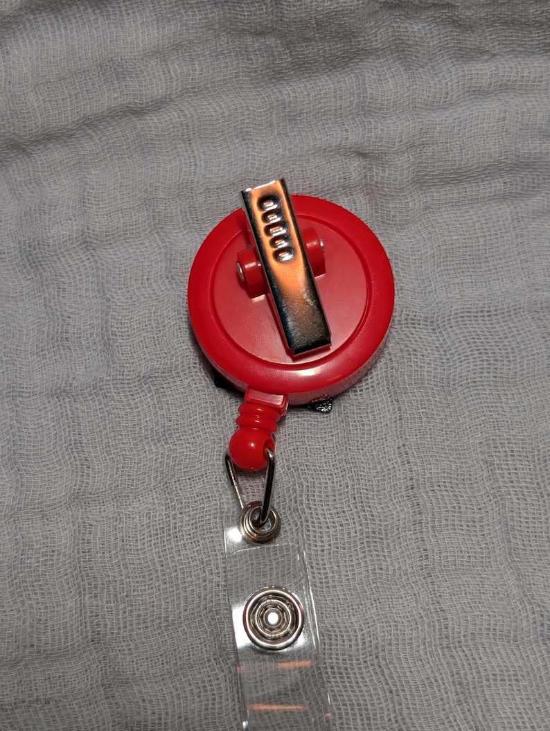 May include: A red retractable badge reel with a silver metal clip and a clear plastic strap. The reel is round and has a small red button. The strap has a metal snap closure.