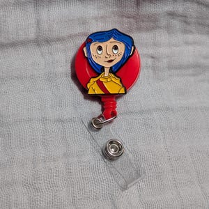 May include: A red and yellow badge reel featuring a cartoon character with blue hair and freckles. The badge reel has a clear plastic strap and a metal clip. The character is wearing a yellow shirt with a red sash.