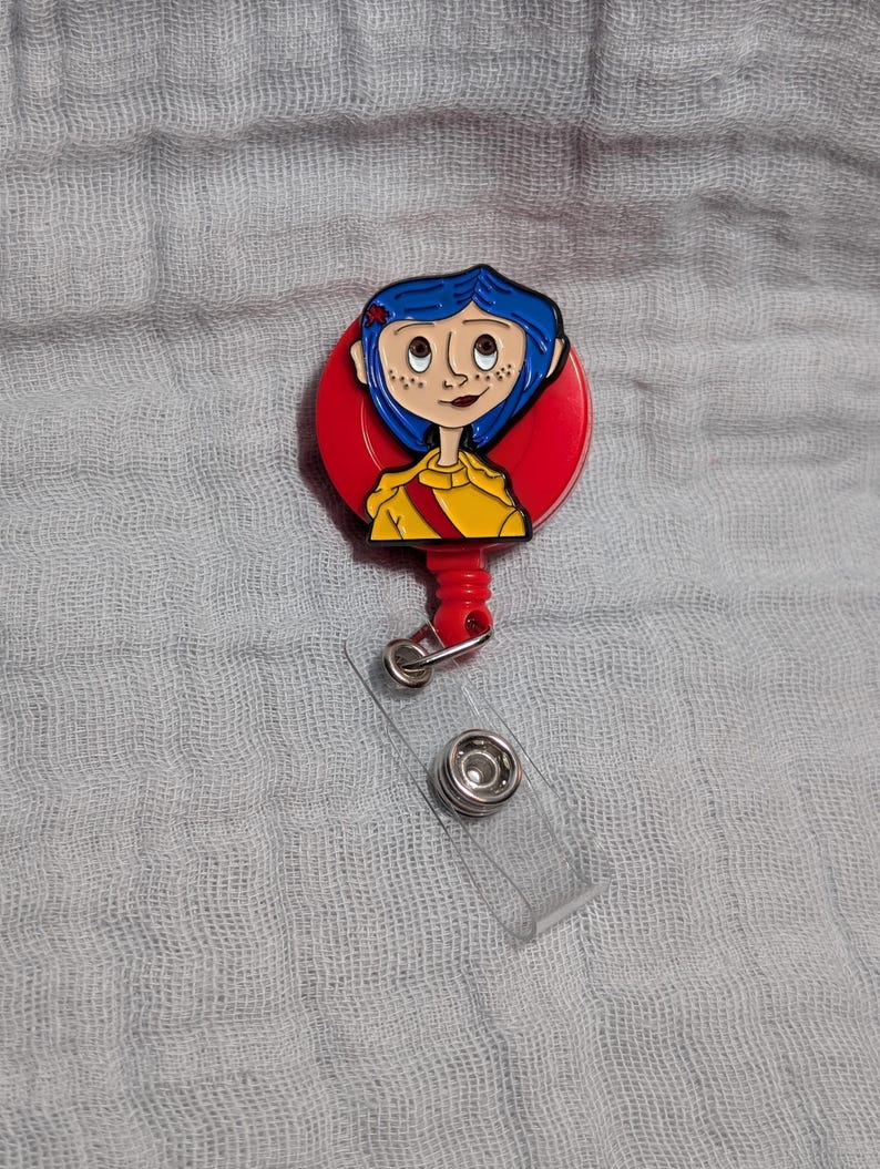 May include: A red and yellow badge reel featuring a cartoon character with blue hair and freckles. The badge reel has a clear plastic strap and a metal clip. The character is wearing a yellow shirt with a red sash.