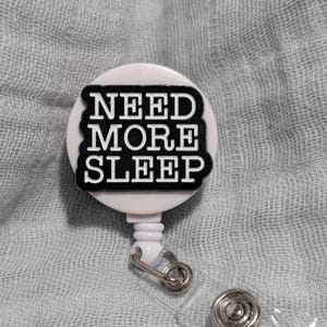 May include: White retractable badge holder with a black and white round button that says "NEED MORE SLEEP".