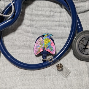 May include: A blue stethoscope with a floral lung-shaped badge attached to a retractable badge reel. The badge is pink, blue, green, and yellow.