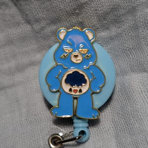 May include: A blue and gold badge reel featuring a cartoon bear with a grumpy expression. The bear has a dark blue belly with a cloud design and three red hearts. The badge reel has a retractable cord and a metal clip.