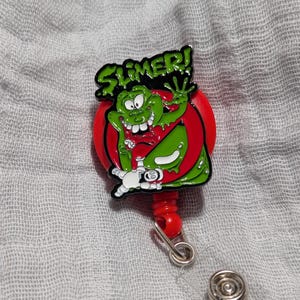 May include: A red and green retractable badge holder with a cartoon character of Slimer from Ghostbusters. The character is green and has a big smile with teeth. The text "SLIMER!" is written in black letters above the character.