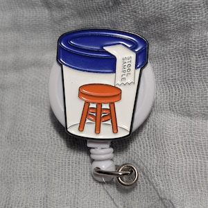 May include: A white badge reel with a cartoon image of a container with a blue lid and an orange stool inside. The text "STOOL SAMPLE" is on a white label. The reel has a clear plastic strap and a metal clip.