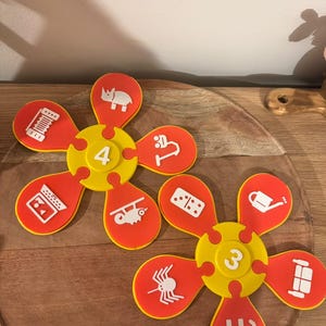 May include: Two flower-shaped educational puzzles made of yellow and orange plastic. One center piece features the number 4 with five orange petals showing white icons of a rhino, microscope, laptop, chameleon, and accordion. The other features the number 3 with petals showing a domino, watering can, sofa, elephant, and spider.