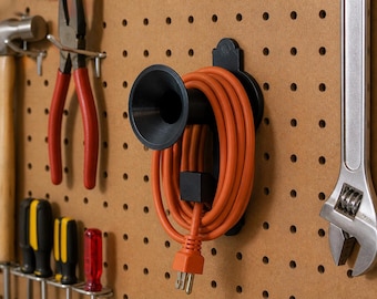 Könnte beinhalten: Ein schwarzer Wand-Kabelhalter aus Kunststoff mit einem trichterförmigen Ende, an dem ein aufgewickeltes, leuchtend oranges Verlängerungskabel hängt. Der Halter ist an einer braunen Lochwand neben verschiedenen Werkzeugen befestigt.