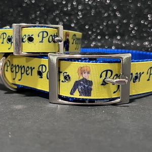 May include: A yellow dog collar with the name "Pepper Potts" printed on it in blue. The collar has a blue interior and a silver buckle. A cartoon image of a person is printed on the collar.