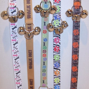 May include: Five dog training bells with different patterned ribbons. The ribbons have paw prints, bones, and the text "Walk Me!" printed on them. The bells are silver and have a ring attached to them.
