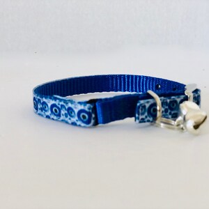 May include: A blue cat collar with a white and blue patterned design and a silver bell. The collar is made of a durable material and features a metal buckle for secure fastening. The bell is small and round, adding a decorative element.