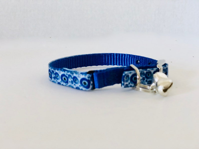 May include: A blue cat collar with a white and blue patterned design and a silver bell. The collar is made of a durable material and features a metal buckle for secure fastening. The bell is small and round, adding a decorative element.