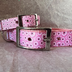 May include: Two pink dog collars with a repeating pattern of white circles and black dots. The collars have silver metal buckles and D-rings. The collars are designed for pets.