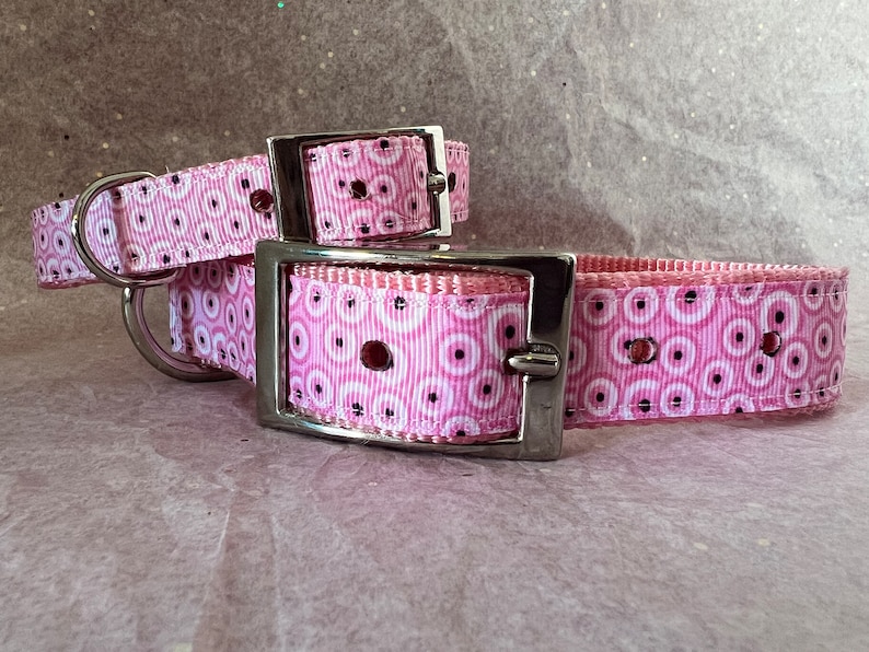 May include: Two pink dog collars with a repeating pattern of white circles and black dots. The collars have silver metal buckles and D-rings. The collars are designed for pets.