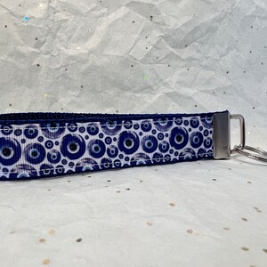 May include: A blue and white wristlet keychain featuring a repeating pattern of blue evil eye symbols. The keychain has a silver-coloured metal clasp and a key ring. The wristlet is made of a sturdy fabric.