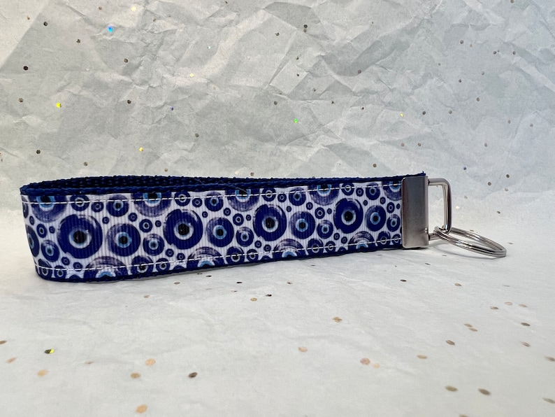 May include: A blue and white wristlet keychain featuring a repeating pattern of blue evil eye symbols. The keychain has a silver-coloured metal clasp and a key ring. The wristlet is made of a sturdy fabric.