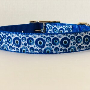 May include: A blue dog collar with a white ribbon patterned with blue and navy blue evil eye symbols. The collar has a silver metal buckle and a blue nylon backing.