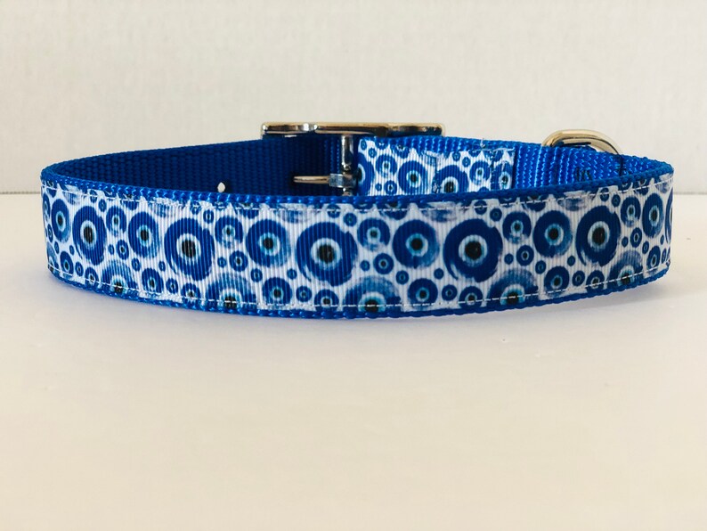 May include: A blue dog collar with a white ribbon patterned with blue and navy blue evil eye symbols. The collar has a silver metal buckle and a blue nylon backing.
