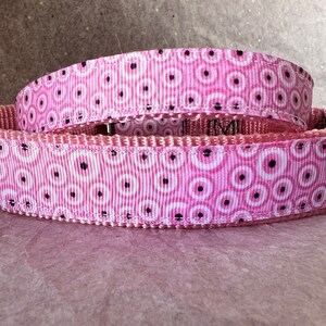 May include: A pink dog collar featuring a repeating pattern of white circles with black centres. The collar is made of a durable, woven material. The background is a light pink colour.