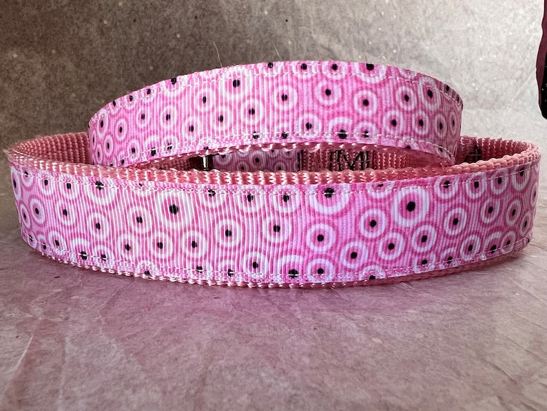 May include: A pink dog collar featuring a repeating pattern of white circles with black centres. The collar is made of a durable, woven material. The background is a light pink colour.