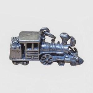 May include: A silver-toned metal charm shaped like a steam locomotive train. The charm features a detailed cabin, a cylindrical boiler, a smokestack, and wheels, with a small circular jump ring attached to the top for hanging purposes.