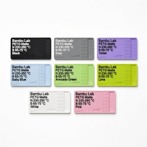 May include: A set of seven rectangular 3D printer filament swatch tags in black, gray, violet, baby blue, avocado green, lime, and white. Each tag features embossed text reading Bambu Lab PETG Matte, temperature settings, and a thickness scale.
