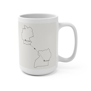 Op de afbeelding: Een witte keramische mok met een zwarte lijntekening van de landkaarten van Duitsland en Oeganda. Een stippellijn geeft een vliegroute aan tussen beide landen met een klein vliegtuigicoon. Stuttgart en Kampala zijn gemarkeerd met rode hartjes.