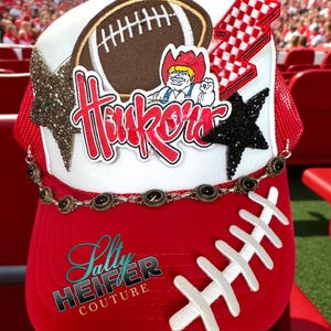 May include: A red and white trucker hat with a football theme. The hat features a brown football patch, a cartoon character, and the word "Huskers" in red. Embellishments include a chain, stars, and a lightning bolt.
