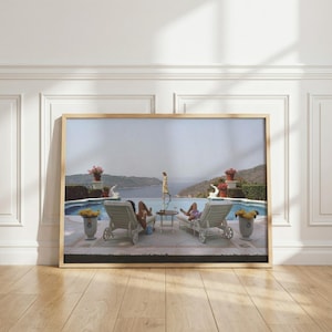 May include: A framed wall art print depicting a swimming pool overlooking a coastal landscape. Two white lounge chairs face the water, with a small table between them. A person in a yellow dress walks near the pool edge, and potted flowers sit on pedestals.