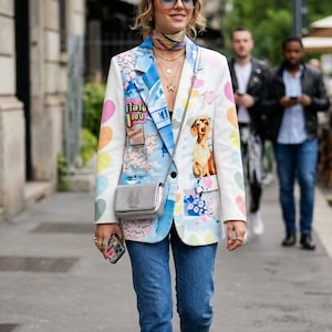 May include: A white blazer featuring a colorful graphic print of hearts, a dog, and the text "Itala Dou" paired with blue jeans, a silver crossbody bag, a beige beret, blue-tinted sunglasses, and white sneakers.