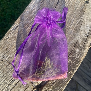 May include: A small, translucent purple organza drawstring pouch featuring a shimmering, sheer fabric texture. The bag is cinched at the top with matching purple ribbon ties and contains small, indistinct organic contents visible inside.