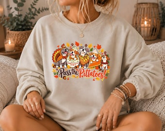 May include: A beige crewneck sweatshirt featuring a colorful graphic of five dogs dressed as Thanksgiving food items, including a turkey and pumpkin pie, with the text Pass the Pittatoes! surrounded by autumn leaves and acorns.