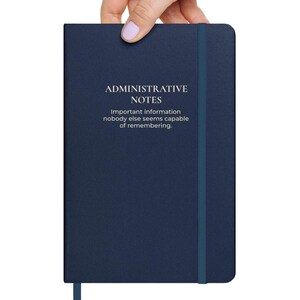 May include: A dark blue hardcover notebook with an elastic closure band. The cover features the white text: ADMINISTRATIVE NOTES, Important information nobody else seems capable of remembering. The notebook has a smooth, solid-colored finish.