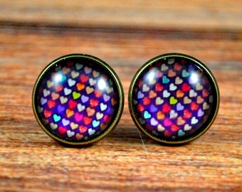 May include: A pair of stud earrings with a round, clear glass design featuring a pattern of colorful hearts on a dark purple background. The earrings have a bronze-colored metal setting.