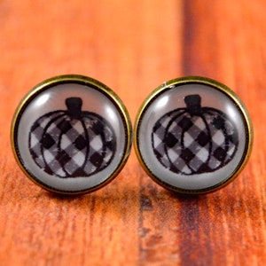 May include: A pair of stud earrings featuring a black and white checkered pumpkin design on a white background. The earrings have a gold-toned metal setting.