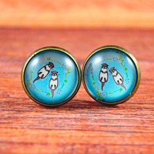 May include: A pair of stud earrings with a blue background and three white and black cats in a circle. The earrings have a bronze metal setting.